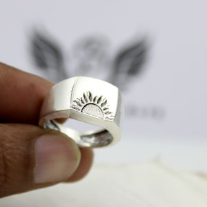 May include: A silver ring with a square-shaped band and a sun design engraved on the top.