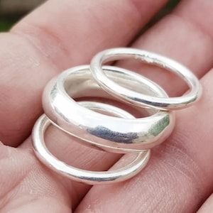 Pode incluir: Um conjunto de três anéis de cor prateada com superfícies lisas e polidas. A coleção inclui uma banda grossa e arredondada posicionada entre duas bandas circulares mais finas e simples, todas com um acabamento metálico reflexivo.