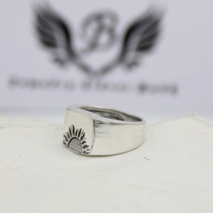 May include: A silver ring with a raised, textured design of a sun on the top. The ring is on a white surface.