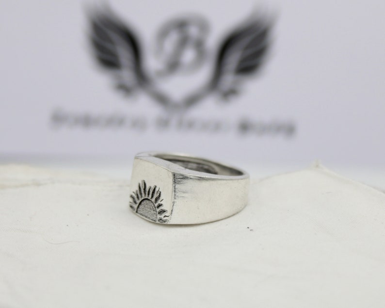May include: A silver ring with a raised, textured design of a sun on the top. The ring is on a white surface.