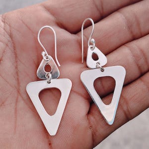 Könnte beinhalten: Ein Paar silberne Ohrringe mit geometrischem Design. Jeder Ohrring hat ein kleines Dreieck über einem größeren Dreieck mit einem Ausschnitt. Die Ohrringe haben einen Hakenverschluss.