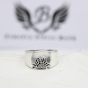 May include: A silver ring with a square-shaped top featuring a raised design of a stylized sun.