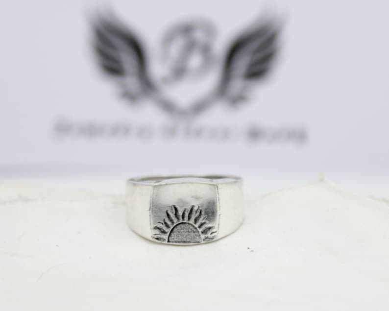 May include: A silver ring with a square-shaped top featuring a raised design of a stylized sun.