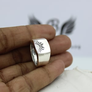 May include: A silver ring with a raised sun design. The ring is worn on a person's finger.