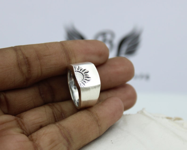 May include: A silver ring with a raised sun design. The ring is worn on a person's finger.