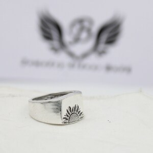 May include: A silver ring with a raised sun design. The ring is a simple band with a square top.