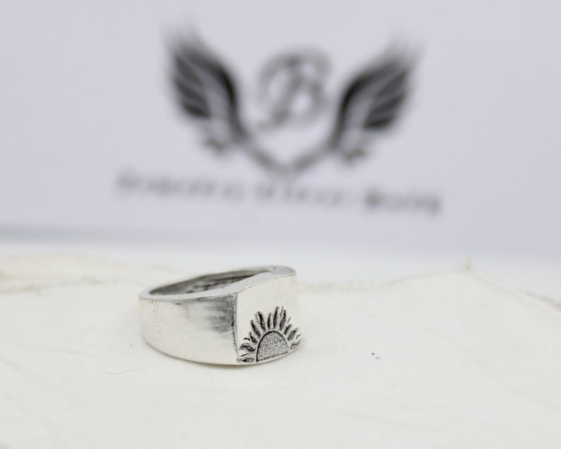 May include: A silver ring with a raised sun design. The ring is a simple band with a square top.