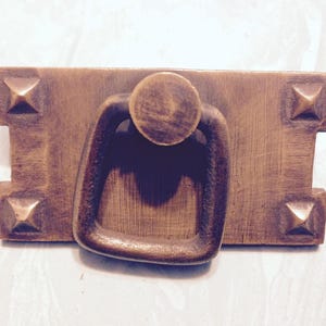 May include: A bronze-toned cabinet pull with a rectangular backplate and a rounded handle. The backplate features four raised, pyramid-shaped accents. The handle is a rounded rectangle shape. The hardware has a vintage aesthetic.