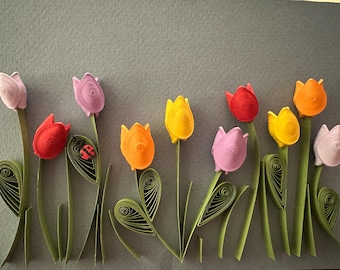 Peut inclure: Une œuvre d'art en papier roulé représentant une rangée de tulipes colorées dans des tons de rose, rouge, orange et jaune. Chaque fleur est faite de bandes de papier enroulées avec des tiges et des feuilles vertes. Une coccinelle est fixée.