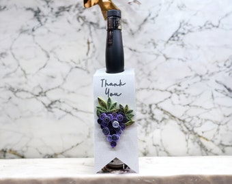 Peut inclure: Une bouteille de vin noire dans une étiquette cadeau blanche avec les mots "Thank You". L'étiquette présente un motif de quilling en papier d'une grappe de raisins violets avec des feuilles vertes. Le fond est un marbre blanc.