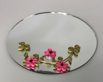 Peut inclure: Un miroir rond avec un bord argenté, décoré de fleurs en papier. Les fleurs sont roses et blanches, avec des feuilles et des tiges vertes. Le miroir est sur une surface blanche.