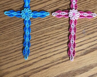Peut inclure: Deux croix en papier faites à la main. L'une est bleue, avec un motif floral bleu clair à l'intersection. L'autre est rose, avec un motif floral blanc à l'intersection. Les deux sont sur une surface en bois.