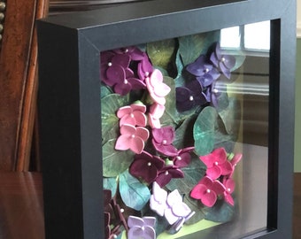 Peut inclure: Une œuvre d'art florale encadrée avec des fleurs d'hortensia roses, violettes et blanches, sur fond de feuilles vertes. L'œuvre est encadrée dans un cadre noir carré avec une façade en verre, créant un effet de profondeur.