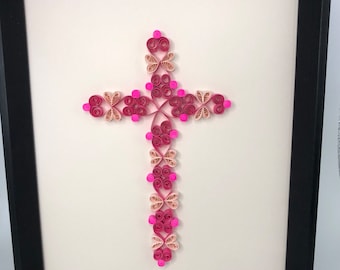 Peut inclure: Une œuvre d'art encadrée représentant une croix en filigrane. La croix est réalisée en papier rose et beige clair, avec un design détaillé et superposé. L'œuvre est placée sur un fond blanc et encadrée dans un cadre noir.