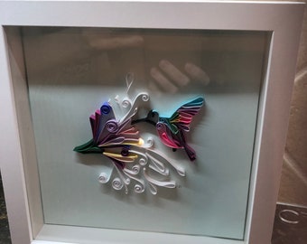 Peut inclure: Une œuvre d'art en quilling encadrée représentant un colibri et des fleurs. Le colibri a des ailes colorées et est positionné près d'une grappe de fleurs. L'œuvre est placée sur un fond bleu clair dans un cadre blanc.