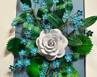 Peut inclure: Une œuvre d'art florale encadrée. Une grande rose grise claire est au centre, entourée de myosotis bleus et de feuilles vertes. L'œuvre est posée sur un fond bleu dans un cadre blanc, créant une composition botanique délicate et détaillée.