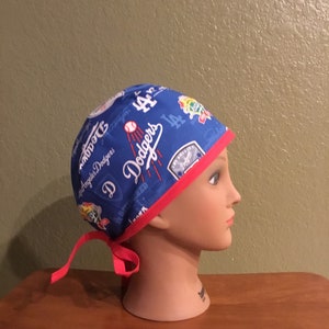May include: Blue and red surgical scrub hat with a Los Angeles Dodgers print. The hat has a red tie and the print features the team logo, baseballs, and the words "Dodgers" and "Los Angeles Dodgers".