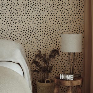 Peut inclure: Papier peint avec un motif répétitif de taches noires irrégulières sur un fond beige. Le mur est situé derrière un tabouret en bois supportant une lampe à abat-jour gris clair et un petit bloc en bois avec le mot HOME imprimé dessus.