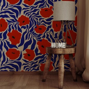 May include: Wallpaper featuring a repeating pattern of vibrant red poppies and deep blue wavy leaves on a cream background. A wooden stool with a lamp and a small white block sign with the word HOME sits in front of the floral wall covering.