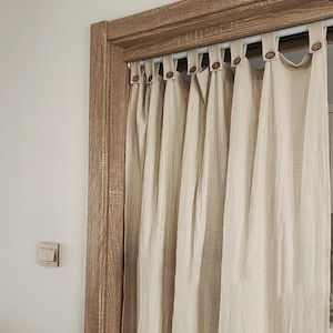 May include: A set of beige linen curtains with wooden button closures hanging from a white rod. The curtains are hung in a doorway with a wooden frame.