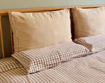 May include: A wooden bed frame featuring two large, solid beige pillows placed against the headboard, with two smaller, rectangular pillows in front. The bed is covered in a light brown and white gingham checkered pattern bedding set.