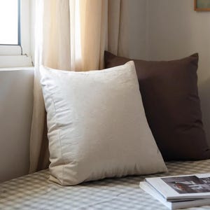 Puede incluir: Un acogedor asiento junto a la ventana con dos cojines cuadrados, uno crema y otro marrón. Una pila de libros descansa sobre el cojín a cuadros. Cortinas suaves de tonos neutros enmarcan la ventana, creando un ambiente relajante.