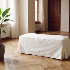 以下が含まれることがあります： プリーツスカートが付いた白い長方形のオットマン。 オットマンは滑らかで明るい色の生地で覆われています。 背景には、寄木張りの床、濃い色の木製キャビネット、シアーなカーテン付きの窓があります。