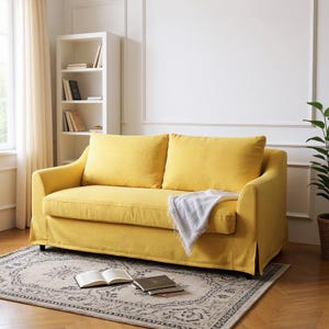 Könnte beinhalten: Ein gelbes Zweisitzer-Sofa mit zwei großen Rückenkissen und einer hellgrauen Decke. Das Sofa steht auf einem gemusterten Teppich, mit einem weißen Bücherregal und einer Pflanze im Hintergrund. Ein offenes Buch liegt auf dem Teppich.