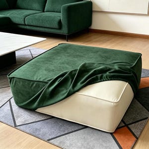 May include: A square ottoman with a dark green velvet top and cream-colored sides. The ottoman is partially covered with a draped green fabric. It sits on a patterned rug, with a green sofa and white coffee table in the background.