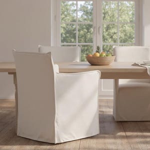 May include: A light-colored wooden dining table paired with three upholstered chairs featuring off-white slipcovers. The table surface holds a wooden bowl filled with various fruits and a small white bowl resting on a light-colored fabric runner.