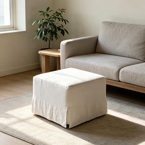 May include: A square, off-white fabric ottoman featuring a slipcover design with a pleated skirt detail. The ottoman has a structured, boxy silhouette and is upholstered in a light-colored, textured textile that sits flat on the floor.