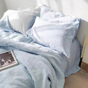 May include: A light blue linen bedding set with a duvet cover, two pillowcases, and a flat sheet. The bedding is arranged on a bed with a wooden floor.
