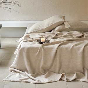 May include: A beige linen bedspread with two beige linen pillows on a bed. A small white candle sits on a round, dark gray tray on the bedspread.