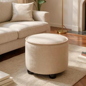 Può includere: Un pouf rotondo color beige con una finitura in tessuto a coste testurizzato e un bordo profilato lungo la circonferenza superiore. L'elemento è sostenuto da quattro piccoli piedini arrotondati color marrone scuro, che formano una base stabile.
