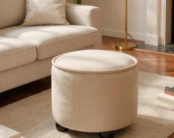 May include: A round, beige ottoman featuring a textured corduroy fabric finish and a piped edge along the top circumference. The piece is supported by four small, dark brown, rounded feet, providing a stable base for this cylindrical furniture item.