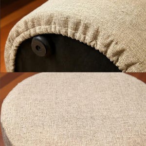 以下が含まれることがあります： ベージュの布張りのスツールのクローズアップ。生地は質感のある外観です。スツールの底は黒で、黒い足が付いています。スツールは丸く、耐久性のある素材で作られているようです。