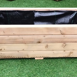 May include: A rectangular wooden planter box with a black liner inside. The planter is made of natural wood and has a rustic look.