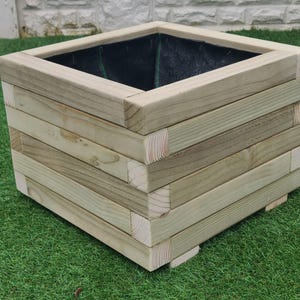 May include: A square wooden planter box with a dark brown liner. The planter is made of natural wood with a light brown finish and has a simple, rustic design.