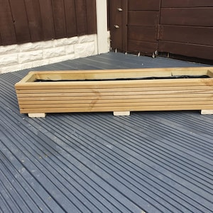 May include: A wooden planter box with a black liner, sitting on a grey composite decking.