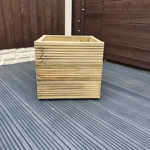 May include: A wooden planter box with a slatted design. The planter is made of natural wood and has a rectangular shape. It is sitting on a grey deck.