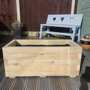 May include: A large wooden planter box with a dark brown liner. The planter is made of natural wood and has a rectangular shape. The planter is sitting on a grey wooden deck.
