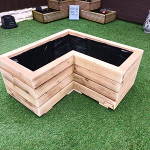May include: An L-shaped wooden planter, constructed from light-coloured timber. The planter box features a black liner and is positioned on green artificial turf. The design incorporates horizontal wooden planks.