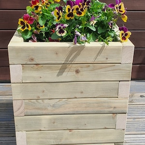 May include: A wooden planter box with a light brown finish. The planter is filled with a variety of colourful flowers, including yellow, purple, and red.