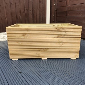 May include: A large, rectangular wooden planter box with a slatted design. The planter is made of natural wood and has a light brown colour. It is sitting on a grey deck.
