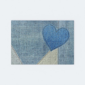 May include: A blue denim heart is sewn onto a patchwork of blue denim fabric. The fabric is cut into jigsaw pieces.