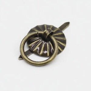 May include: A bronze-toned metal drawer pull featuring a circular ring attached to a round, fluted backplate. The hardware has a weathered, antique finish with a pointed metal prong extending from the back of the circular base component.