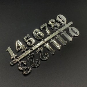 May include: A set of silver-coloured plastic numbers, 1 through 9 and 0, arranged in two rows on a silver strip. The numbers are slightly raised and have a glossy finish.