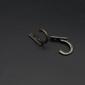 Könnte beinhalten: Zwei antike, bronzefarbene Metallhaken. Jeder Haken hat eine gebogene, abgerundete Form mit einer flachen, rechteckigen Basis. Die Haken sind zum Aufhängen von Gegenständen bestimmt und haben eine Vintage-Ästhetik. Der Hintergrund ist dunkel.