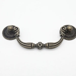 Peut inclure: Poignée de meuble en bronze antique avec des détails ornés et un bouton décoratif à chaque extrémité.