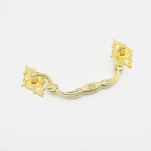 Può includere: Tiramaniglia per cassetto in metallo dorato con dettagli decorativi. La maniglia ha una forma curva e due estremità decorative.